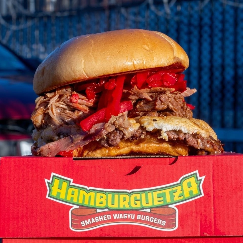 Oversized smashed Wagyu burger topped with pulled meat and red peppers, displayed on a red Hamburguetza box.